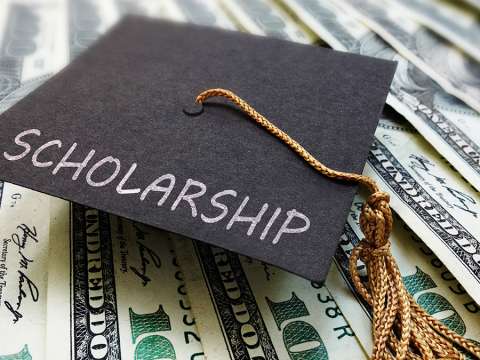 A graduation cap with the word scholarship written on it, sits on top of one-hundred dollar bills.