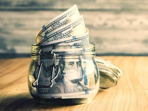 An open jar on top of a wooden table filled with one-hundred dollar bills.