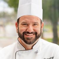 Chef Travis smiling, wearing a white chef's hat and chef's coat.