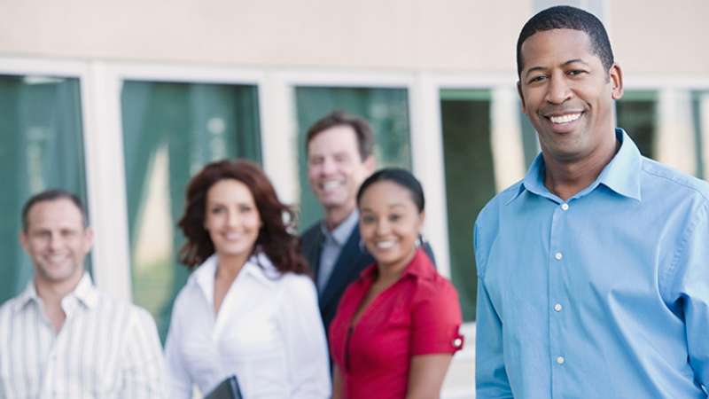 Business people standing outside a building, with one in the foreground, 4 in the background.