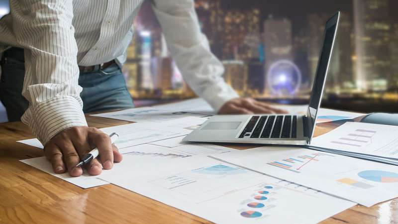 A man is standing at his desk with a laptop looking over charts layed out on his desk