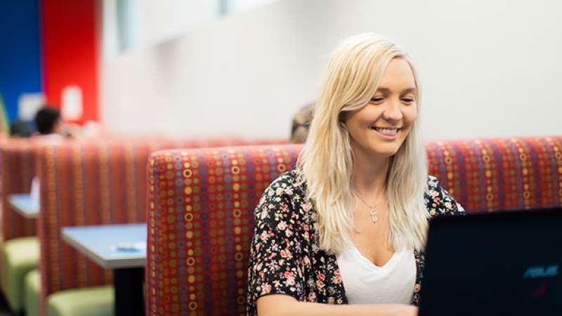 A student is sitting in the NTC cafeteria on her lapton with a confident smile