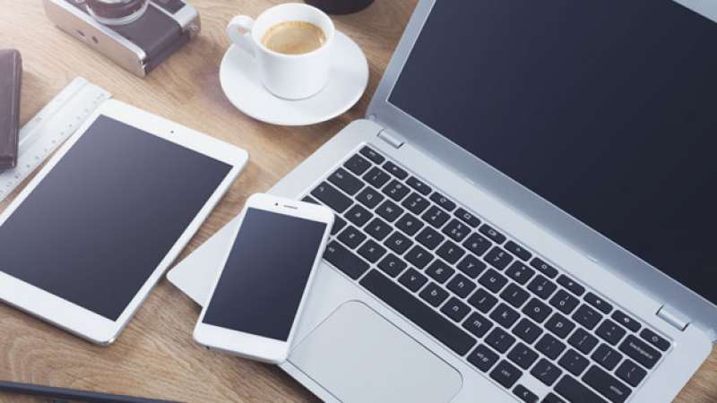A smartphone, tablet, and laptop are set out on a desk next to a camera and a cup of coffee