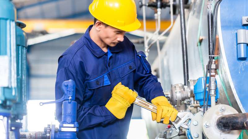 A student wearing a blue jumpsuit and a yellow hardhat is tightening a bolt with a wrench