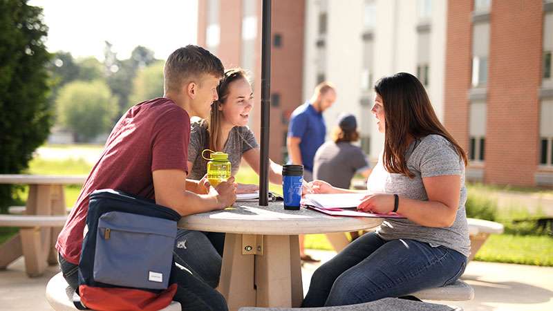 Three students are sitting together working on homework and having a conversation with each other