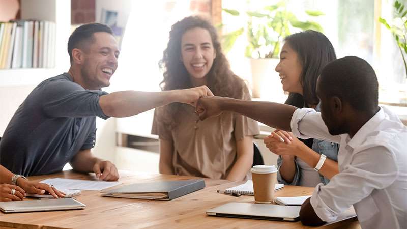 Several people sit at a table having a conversation with coffee. Two men smile and reach across the table to bump knuckles.