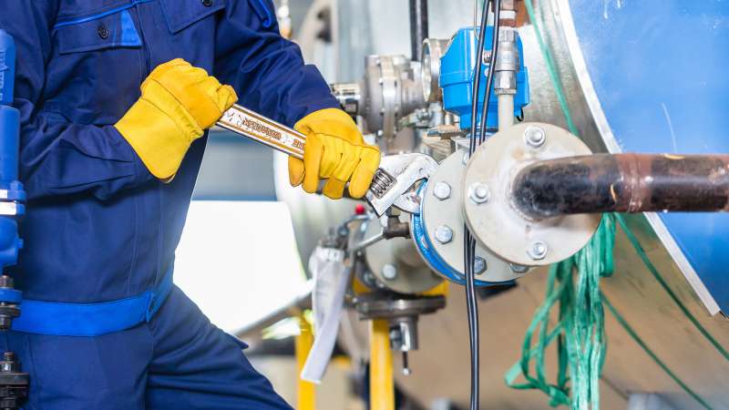 A worker in a blue coverall and orange gloves is using a wrench on large pipes.