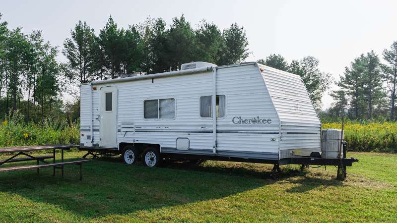 A white Travel Trailer is set up next to a family picnic table on a flat grassy area next to trees and bushes.