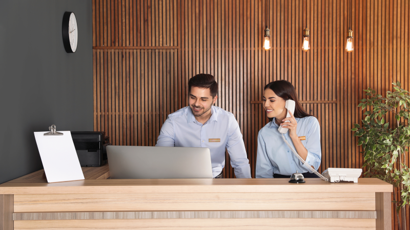 Two front desk employees working at a hotel. One employee is working on a computer and the other is talking on the phone.