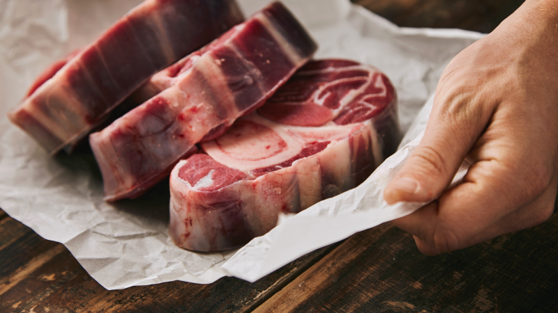 A hand holding three uncooked steaks on top of butchery paper above a wood surface.