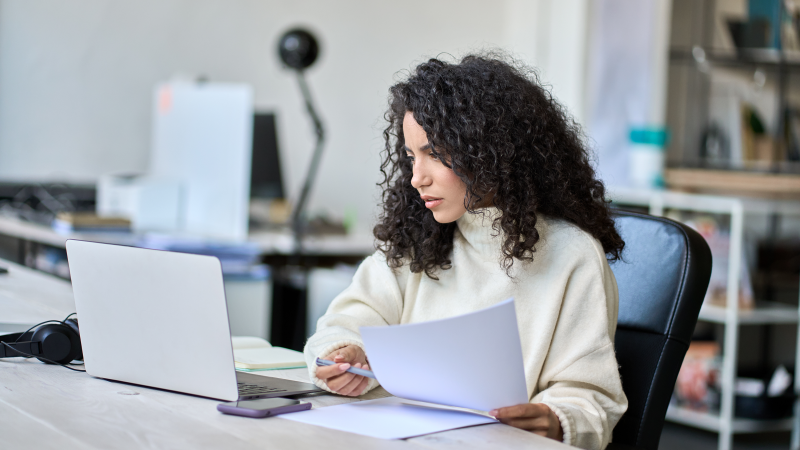 A woman holds some papers in one hand and a pen in the other, as she stares intently at her laptop screen.