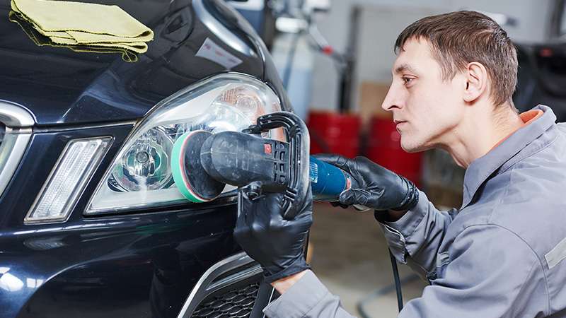 An auto repair person is using a tool to polish the headlight of a vehicle.
