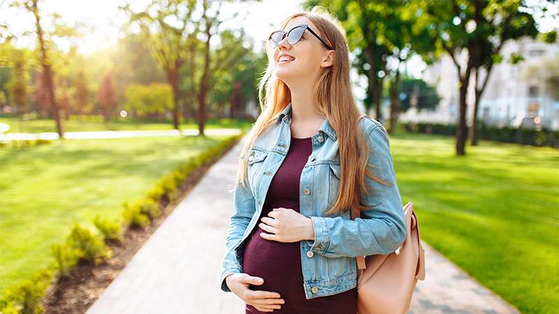 A young pregnant woman walks down the sidewalk holding her belly with a smile on her face.