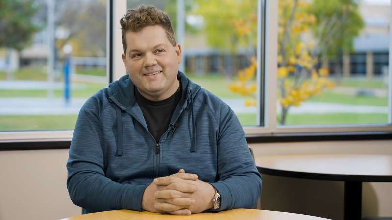 Richard Frahm, a Plumbing Apprentice, sitting in front of several windows with his hands folded on the table in front of him, and smiling while talking about his apprenticeship experience.