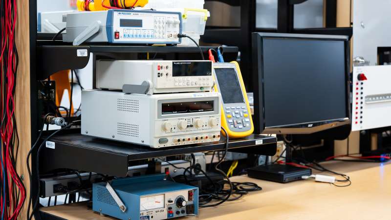 A workstation in an electronics lab featuring various test equipment. Visible are a BK Precision function generator, a Fluke digital multimeter, a dual power supply, and a vintage analog meter device on a shelving unit. There is also a monitor and cables organized around the setup. The workspace appears well-equipped for electronic testing and diagnostics.