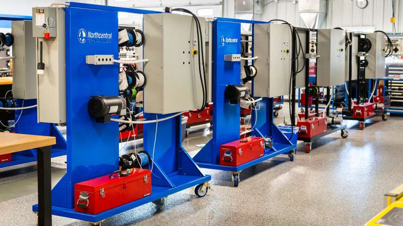 The mechanical systems area with multiple blue training stations on wheels, each equipped with control panels, motors, and spooled cables. The stations are labeled “Northcentral Technical College” and include red toolboxes mounted at the base. The lab is brightly lit, with a polished floor and industrial equipment in the background.
