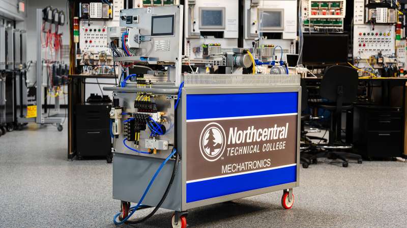 A mobile mechatronics station at Northcentral Technical College, featuring a touchscreen, wiring, pneumatics, and motors. Background shows training stations with control panels in a hands-on learning lab.