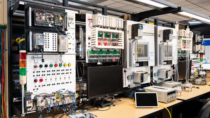 A lab workstation with multiple programmable logic controller (PLC) panels, touchscreens, wiring setups, and indicator lights. The desk features monitors, wiring modules, and testing equipment, creating a hands-on learning environment for automation and control systems.