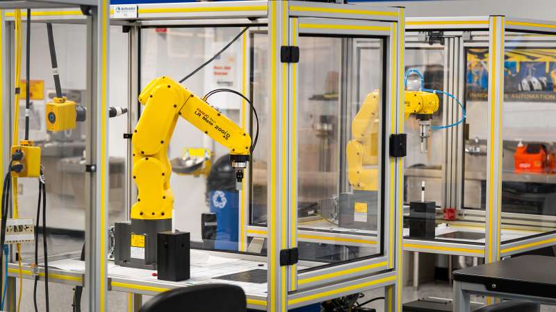 A robotics training setup featuring yellow FANUC LR Mate 200iD robotic arms enclosed in safety cages with clear panels and aluminum frames. The robots are equipped with end-effectors, and the lab environment emphasizes industrial automation and precision training.