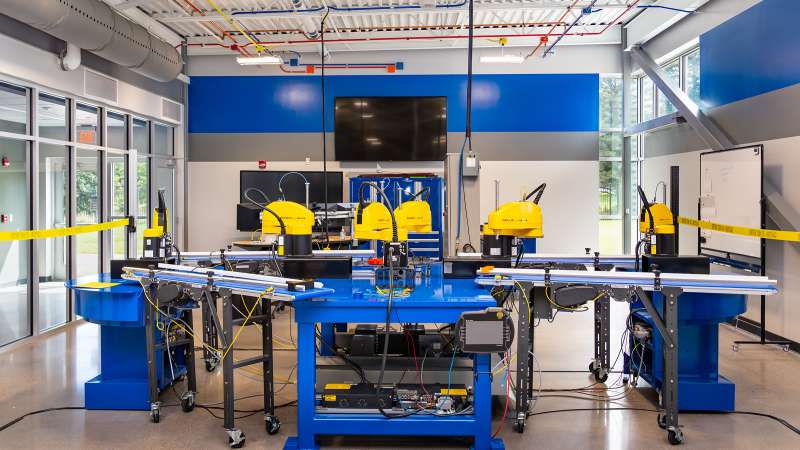 The Industry 4.0 Smart Manufacturing Lab featuring four yellow FANUC SCARA robots on a blue automation cell with conveyor belts. Overhead cables and a central monitor highlight advanced automation and industrial training in a modern, well-lit workspace.
