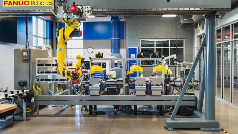 A FANUC Robotics “lights-out” manufacturing system featuring a large robotic arm mounted on a gantry and several yellow SCARA robots working on conveyor-based stations. The lab showcases advanced automation with a focus on precision and efficiency in a clean, modern environment.