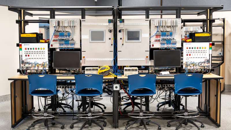 A workstation in the Mechatronics & Fluid Power Lab with two training panels equipped with programmable logic controllers (PLCs), wiring modules, touchscreens, and test equipment. Blue chairs and monitors are positioned for hands-on apprentice training in industrial automation.