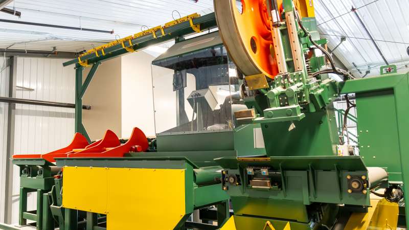 Backside view of the head rig with large orange flywheel, control booth, and red log clamps, part of the advanced sawmill equipment at the training facility.