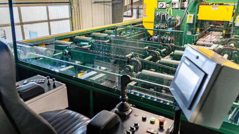 Interior view from a control cab overlooking conveyors and sawmill machinery, featuring a joystick control system, touchscreen, and safety glass window.