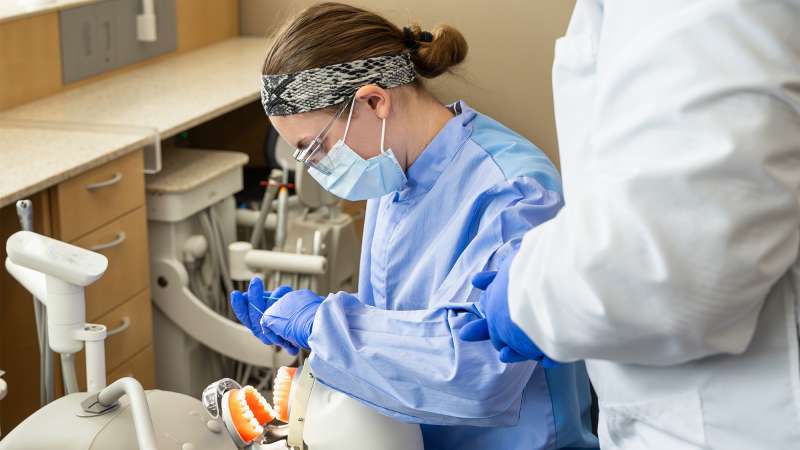 Dental student in blue scrubs practicing on a mannequin, wearing mask and gloves in a clinical setting.