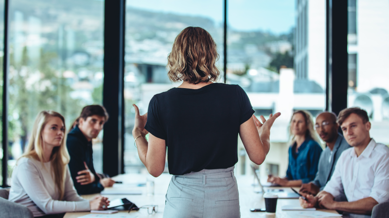 A woman stands with her back to the camera, speaking to a group of colleagues seated around a conference table in a modern office with large windows.