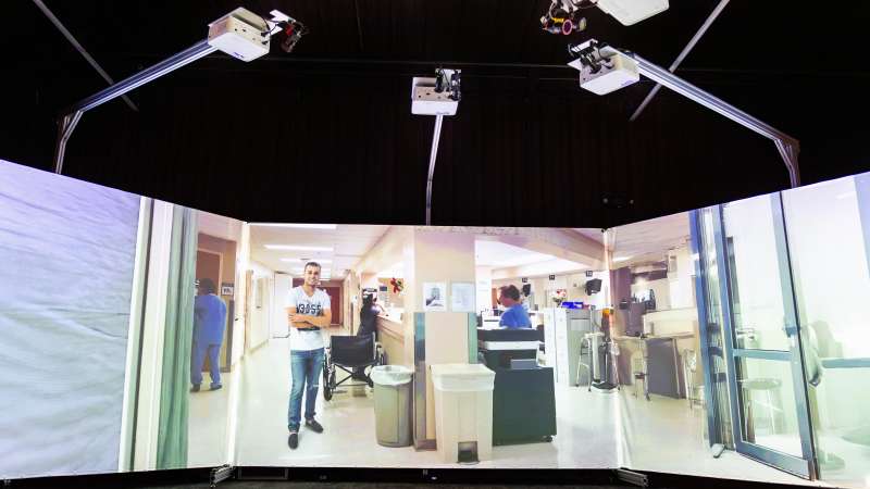 A simulated hospital scene is projected across curved screens in the Community Safety Simulation Center, showing a man standing near a wheelchair in a busy medical facility.