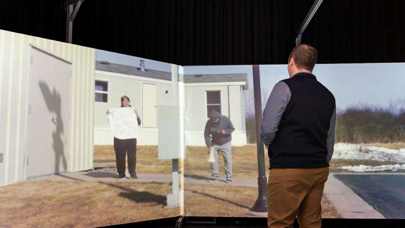 A man takes part in a protest simulation in the Community Safety Simulation Center, where projected individuals stand outside with a megaphone and a sign reading “Turn to the TRUTH!” in a residential setting.