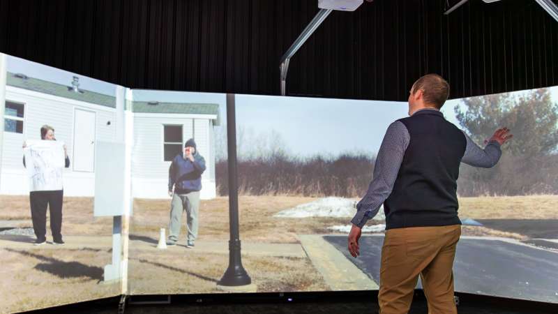 A man interacts with a large, curved screen in the Community Safety Simulation Center, participating in a virtual protest scenario with projected individuals holding signs and filming.