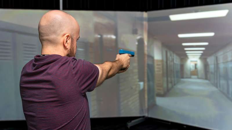 A man participates in an active shooter simulation set in a school hallway, aiming a training weapon at a projected scenario within the Community Safety Simulation Center.
