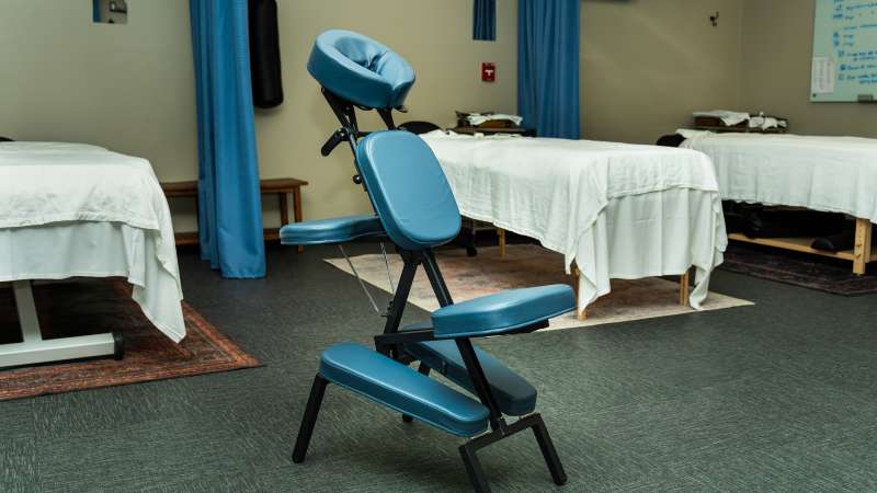 A blue padded massage chair sits in the center of a tranquil room with curtained massage tables and soft lighting, designed for therapeutic treatments.