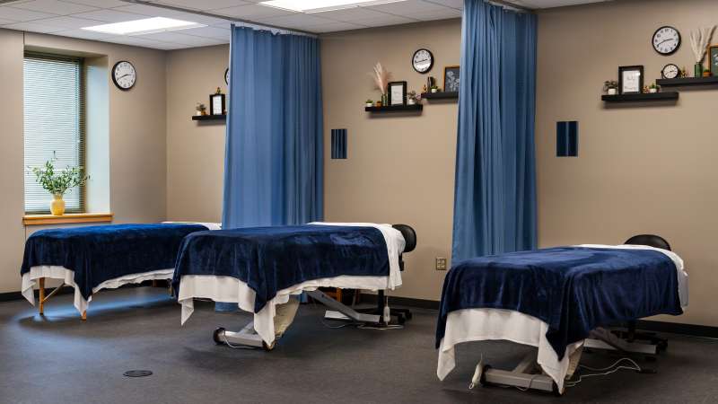A quiet massage therapy room with several massage tables covered in navy blue blankets, separated by blue privacy curtains, and soft natural light.