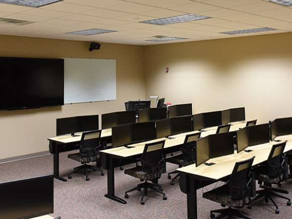 A computer lab classroom setup with several rows of tables with computers, along with an instructor's stand and a TV mounted to the wall.