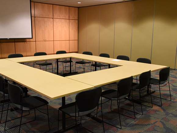 A large training room with the wall closed, dividing the training room into two. Tables are set up in a square pattern, with 4 chairs on each side.