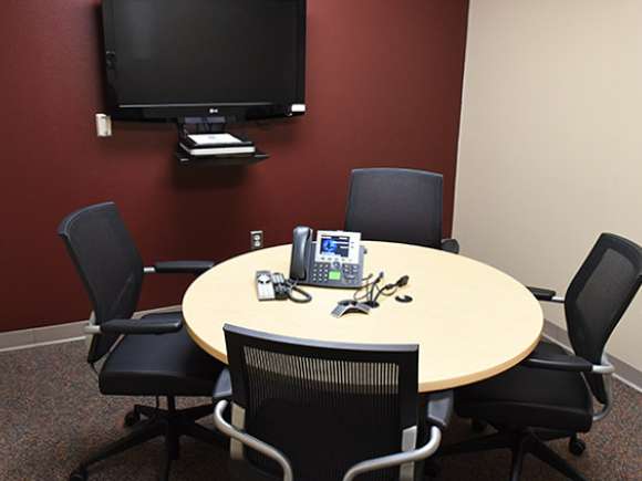 A small IVC room, with four chairs around a small circular table. A TV screen is mounted to one of the walls, and a conference phone sits on the table.
