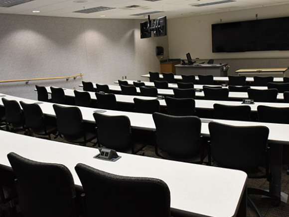 A tiered training room with several rows of tables and chairs in a tiered level format. Three large TV screens/monitors are mounted to the front wall side by side.