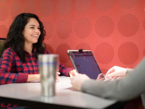 A student sits in a booth near the Campus Cafe, across from another individual, who is in the foreground, just off camera.