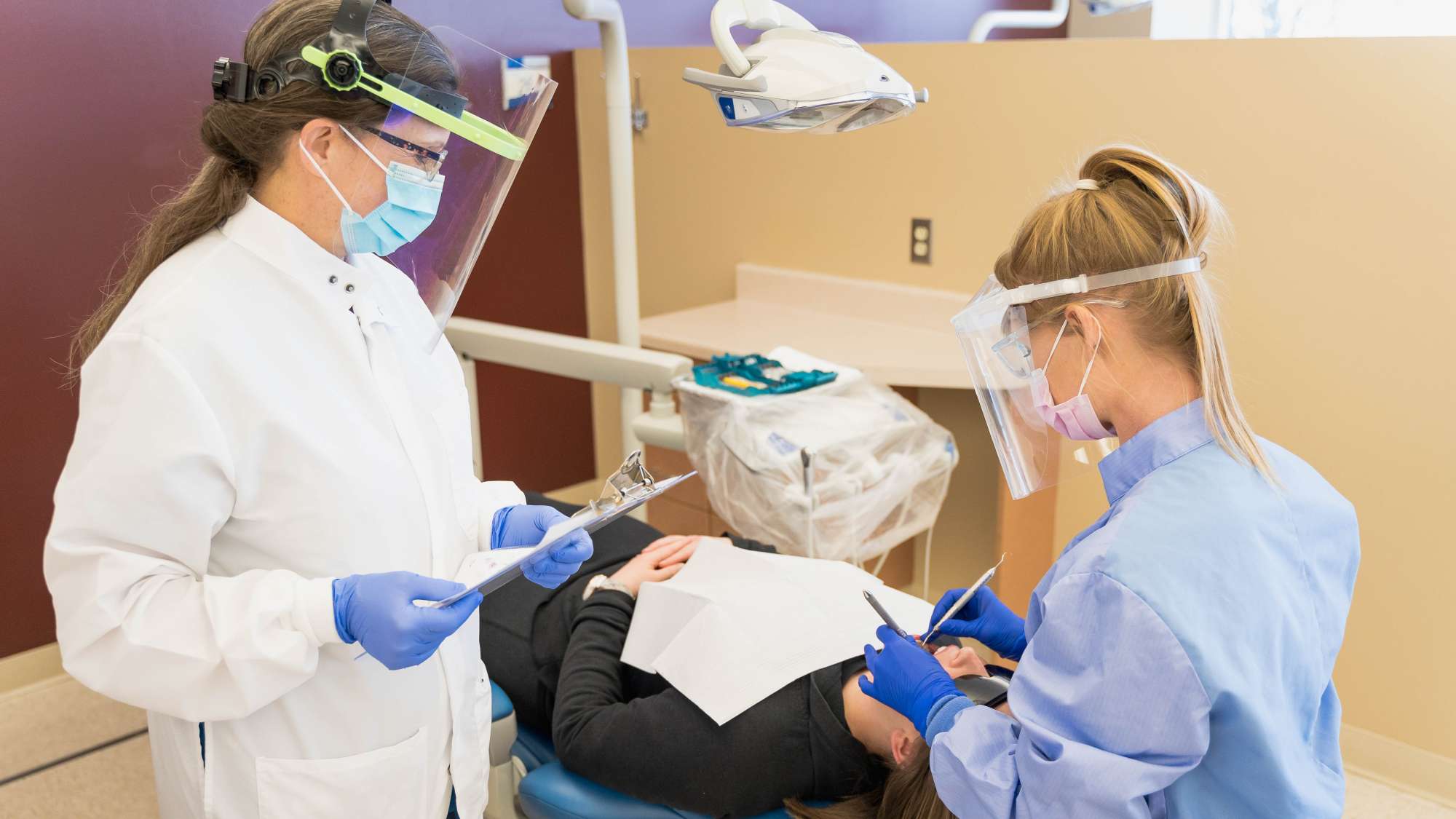 A dental hygienist performs an oral cleaning on a patient while a dentist observes her work with a clipboard in hand.