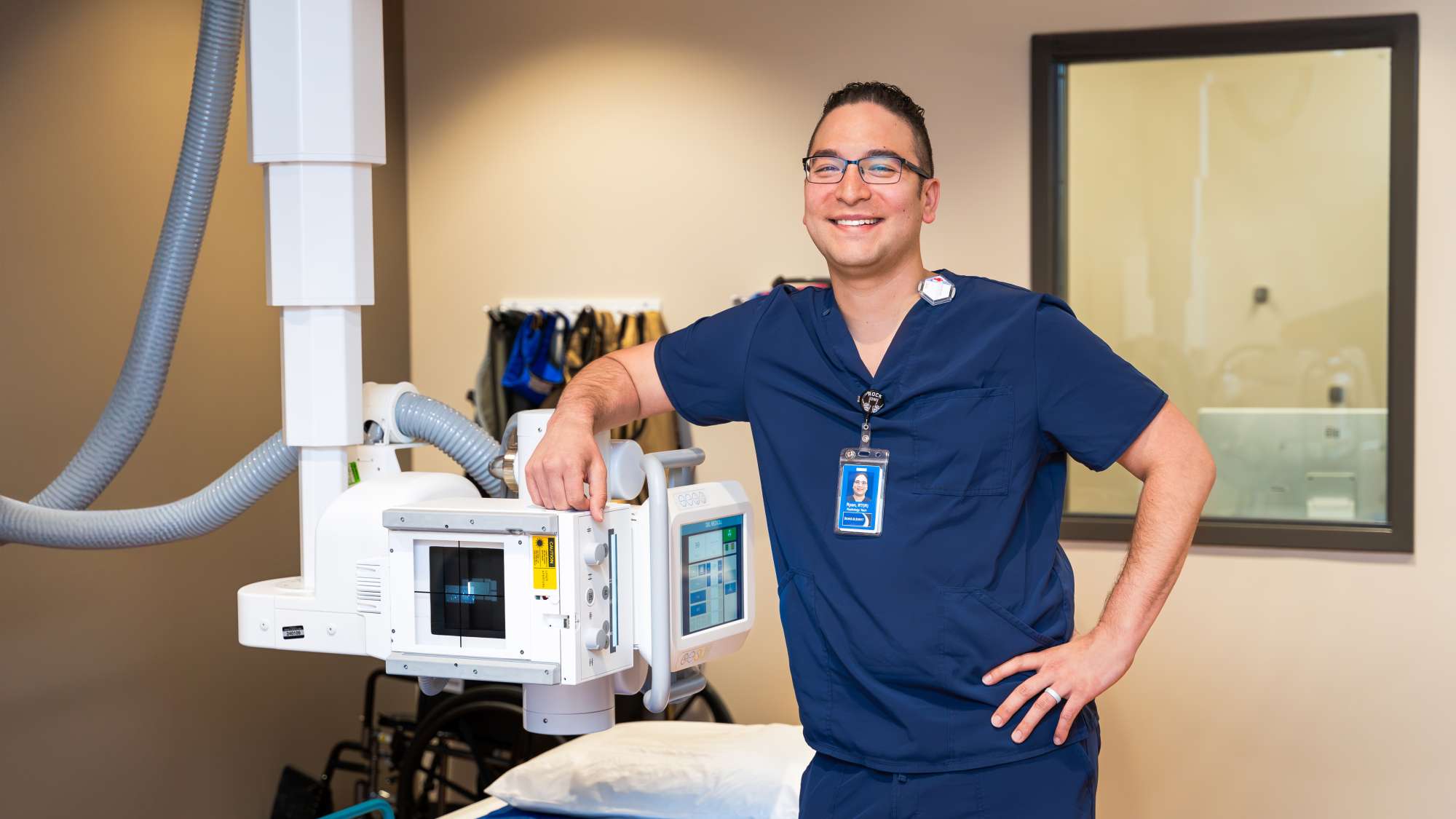 Ryan Whitcraft standing in a radiology room while wearing scrubs, with one arm propped up on radiology equipment that hangs from the ceiling. 