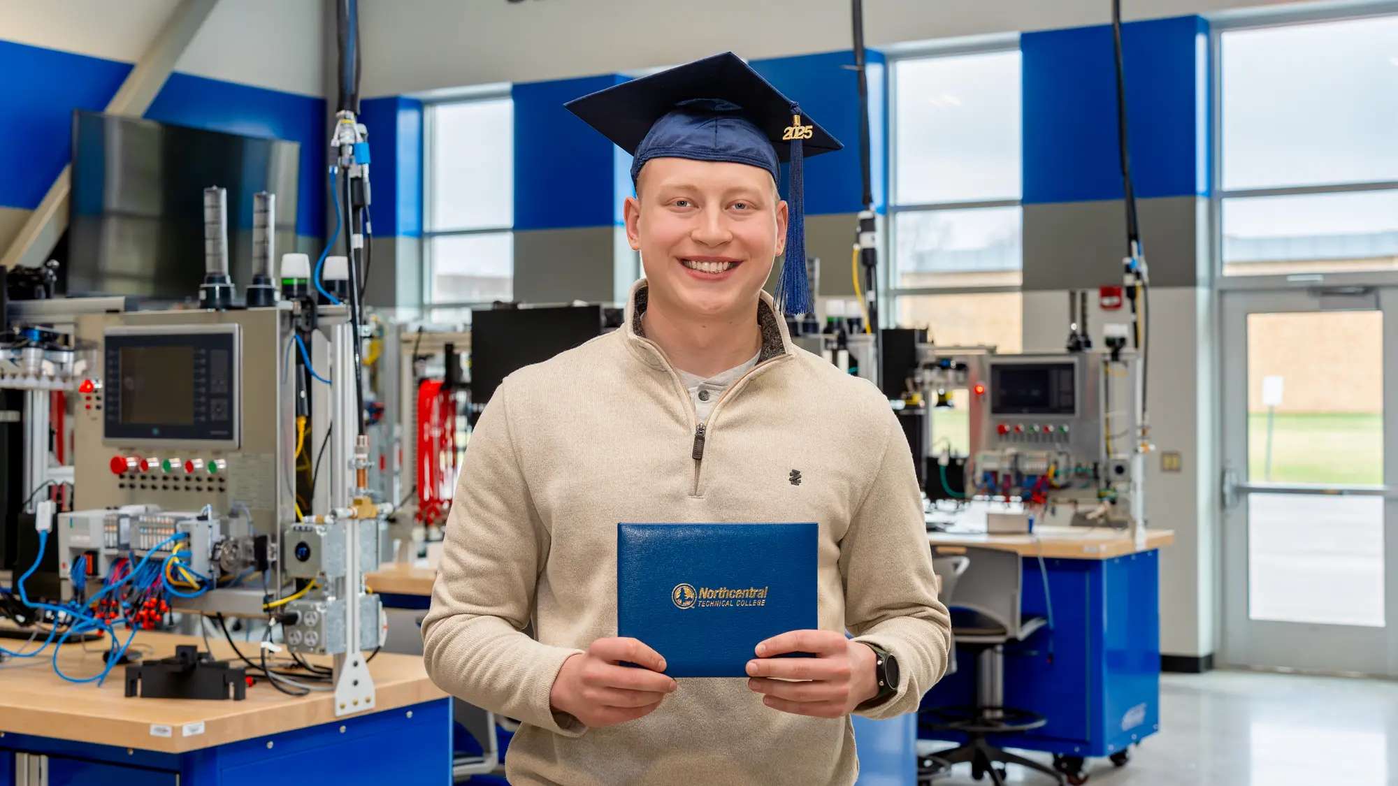 Ethan Sondelski smiling while wearing his graduation cap and holding his diploma while standing inside the Industry 4.0 Smart Manufacturing Lab