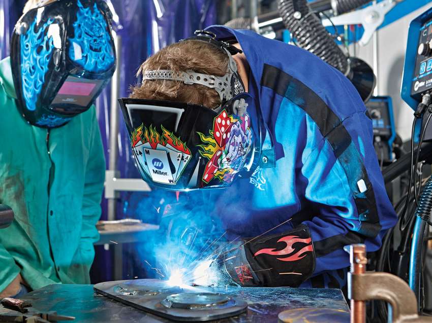 A student is welding medal together as an intern watches. Bright sparks shoot out from the objects being welded together