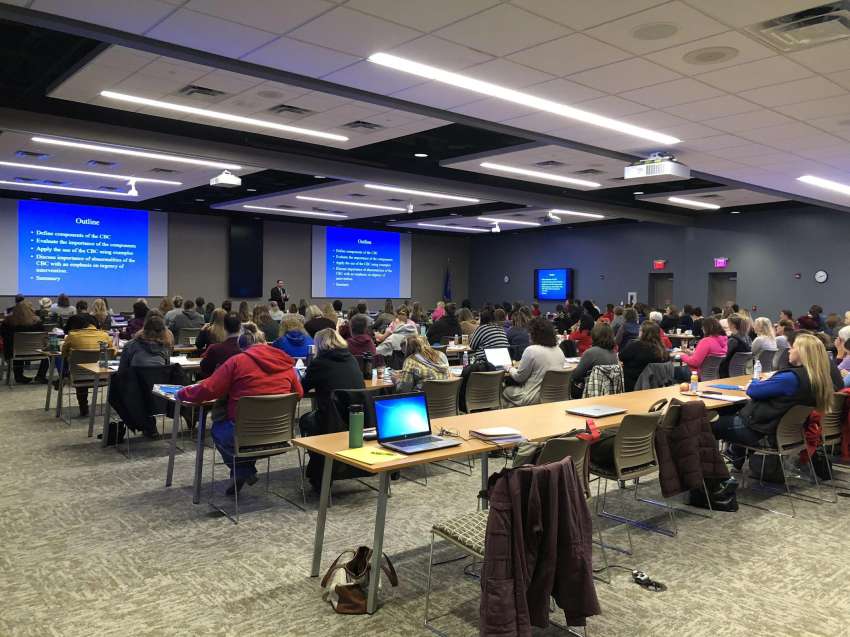 View from the back of the professional conference center with a speaker addressing conference attendees at the front of the room.