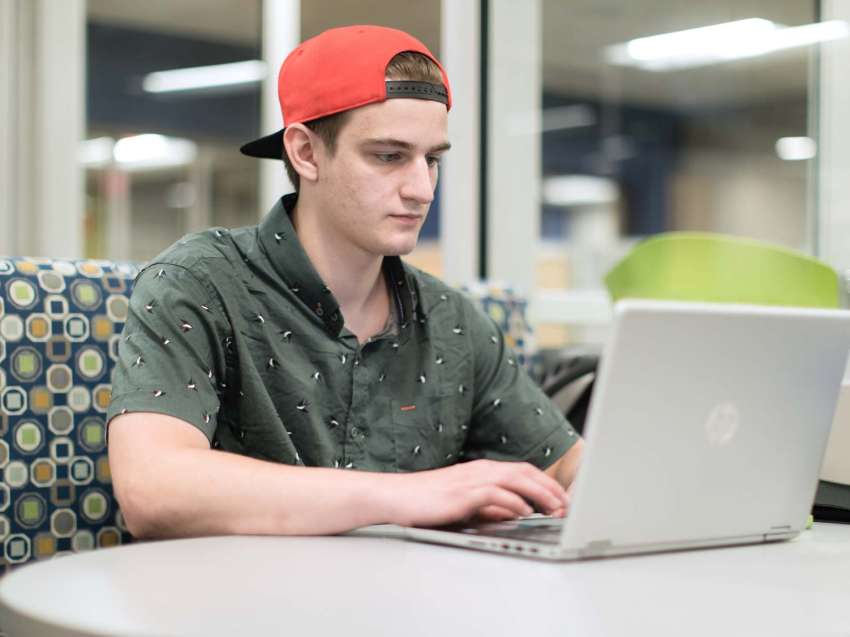 Student sitting in a booth, typing on his laptop, which sits on the table in front of him.