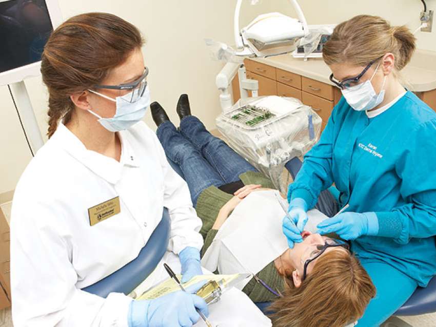 Two dentists stand over a patient inspecting her teeth with dentist tools