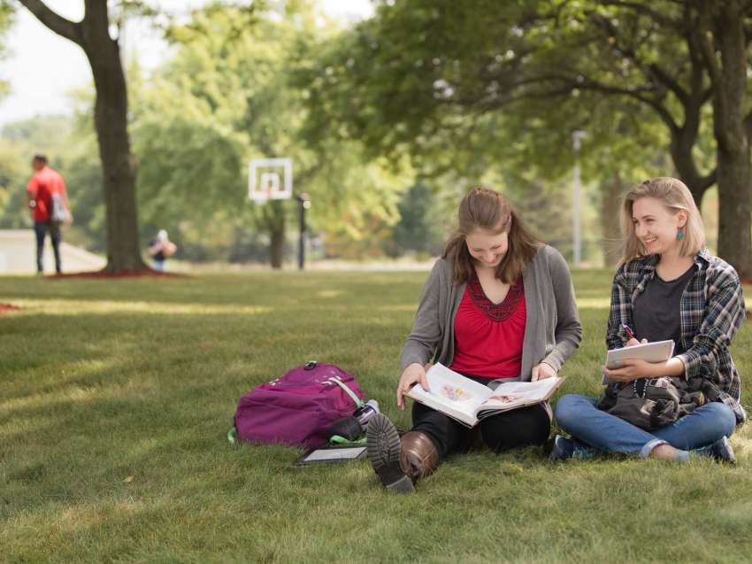 Two students sitting on the grass in the courtyard of the Wausau Campus, as other students are seen in the background.