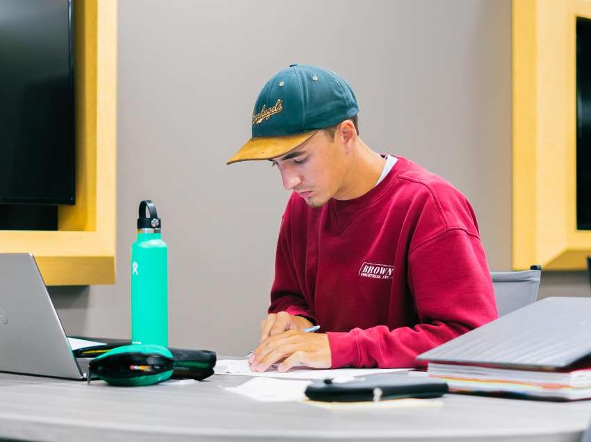 A student working with pencil in hand, an open laptop, papers, and a binder in front of him, at a tabletop in the STEM Center.